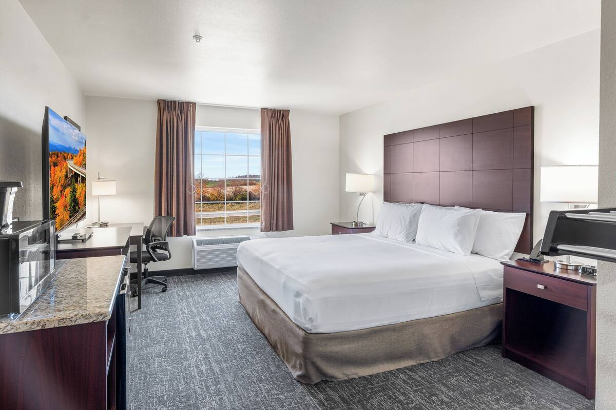 Quarto de hotel com uma cama grande, uma escrivaninha, uma cadeira, uma televisão e uma janela com cortinas.