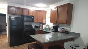 A kitchen with wooden cabinets, a black refrigerator, a microwave, a stove, and a coffee maker. There are two stools and a small table with a remote control. - Cozy 1-bdrm/1 bath, desk, grill, deck, laundry (Phenix City)