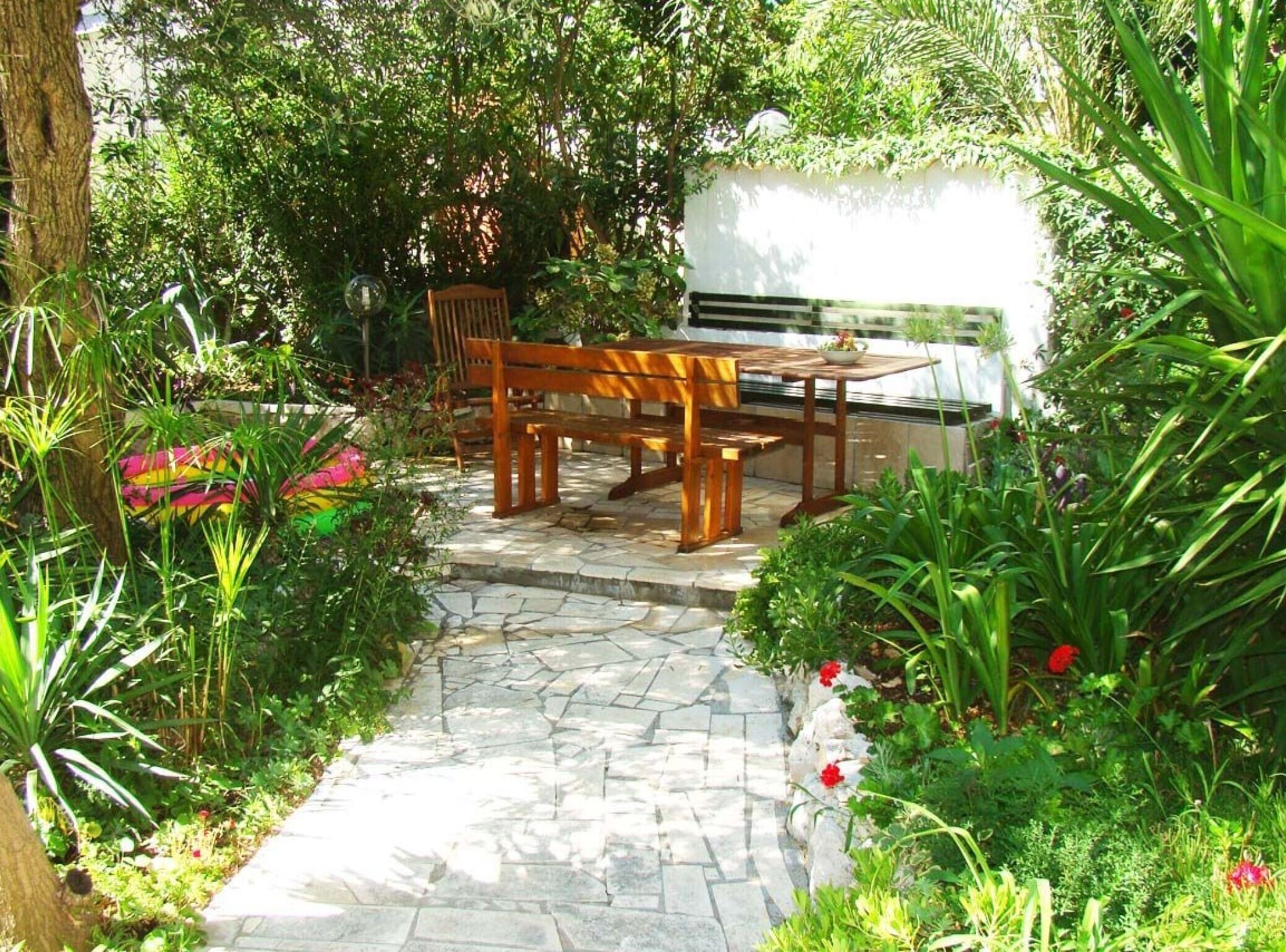 Un chemin de pierres mène à un banc et une table en bois, installés dans un jardin luxuriant où poussent différentes plantes et fleurs.