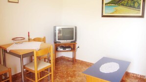 Una stanza modesta con un tavolo in legno, una televisione su un mobile, un quadro incorniciato appeso alla parete e un pavimento piastrellato.