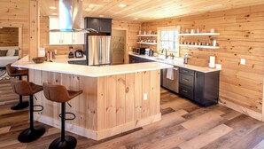 A modern kitchen with wooden paneling, a central island with bar stools, stainless steel appliances, and a window overlooking the outdoors. - Moksha Retreat - A-Frame|Mtn Views|Hot Tub|King Ensuites (Black Mountain)