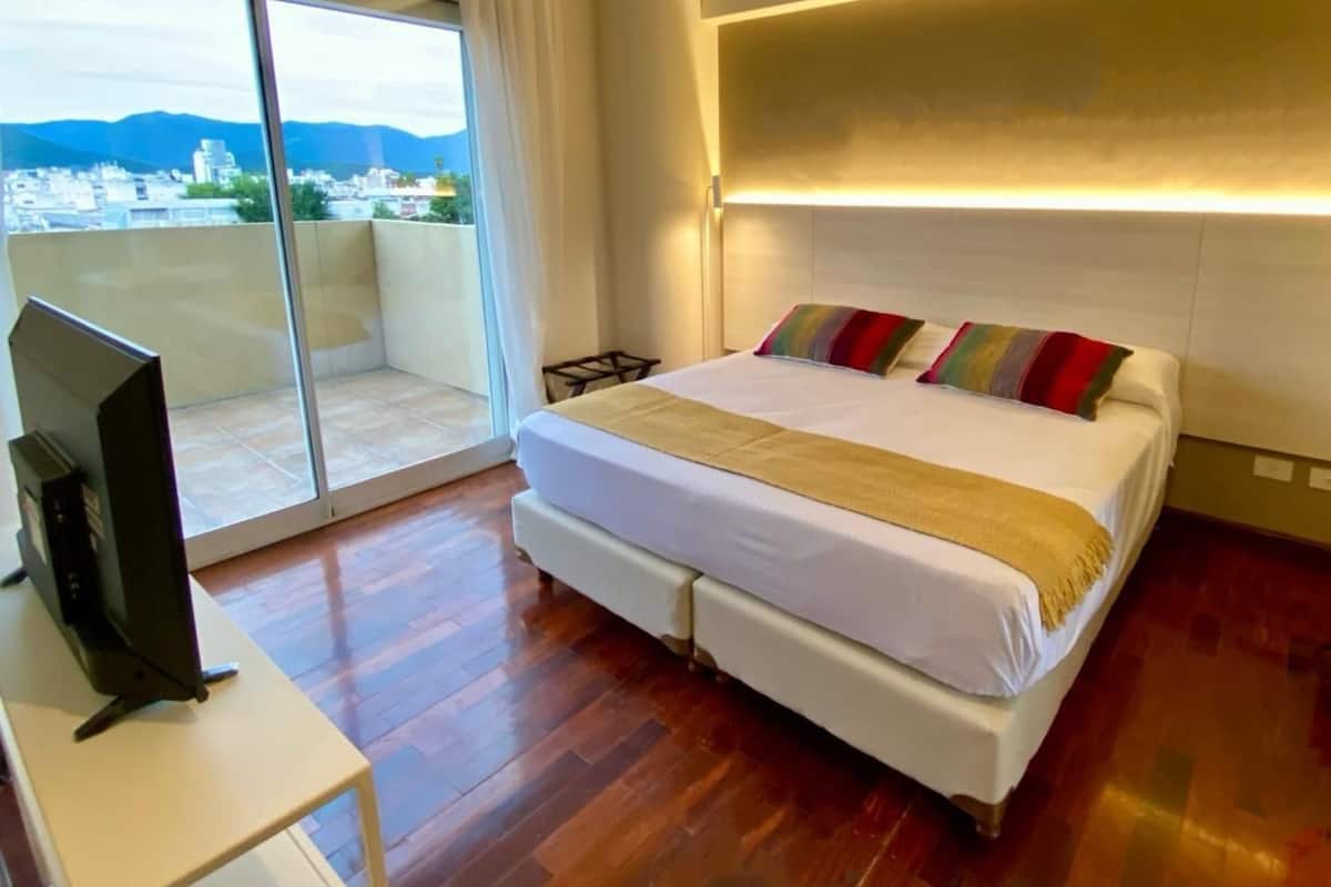 Habitación de hotel con una cama grande, un televisor sobre un mueble bajo y un balcón con vistas a la ciudad.