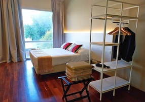 Un dormitorio con cama, una mesita de noche con una maleta, un armario y una ventana con cortinas.