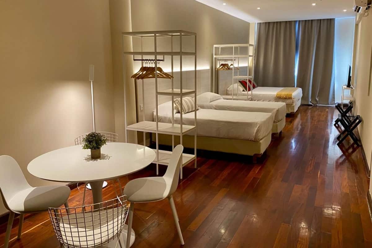 Una habitación de hotel moderna con suelo de madera, una mesa redonda de comedor con sillas, un perchero, dos camas con ropa blanca y un televisor sobre un mueble.