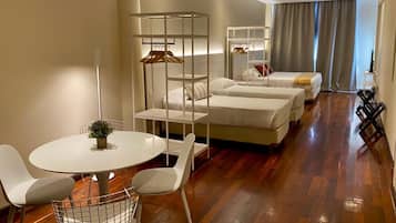 A modern hotel room with a wooden floor, a round dining table, white chairs, a hanging coat rack, and a bed with a white headboard.