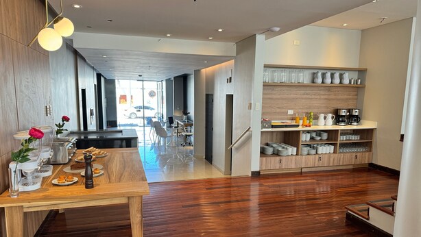 A modern kitchen with wooden flooring, a dining area with a table set for two, and a breakfast bar with coffee and pastries. - Roomie Salta by DOT Suites (Salta)