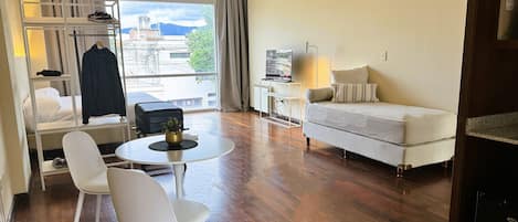 Quarto de hotel com cama, sofá, televisão, mesa com cadeira, escrivaninha e janela com vista para edifícios e montanhas.