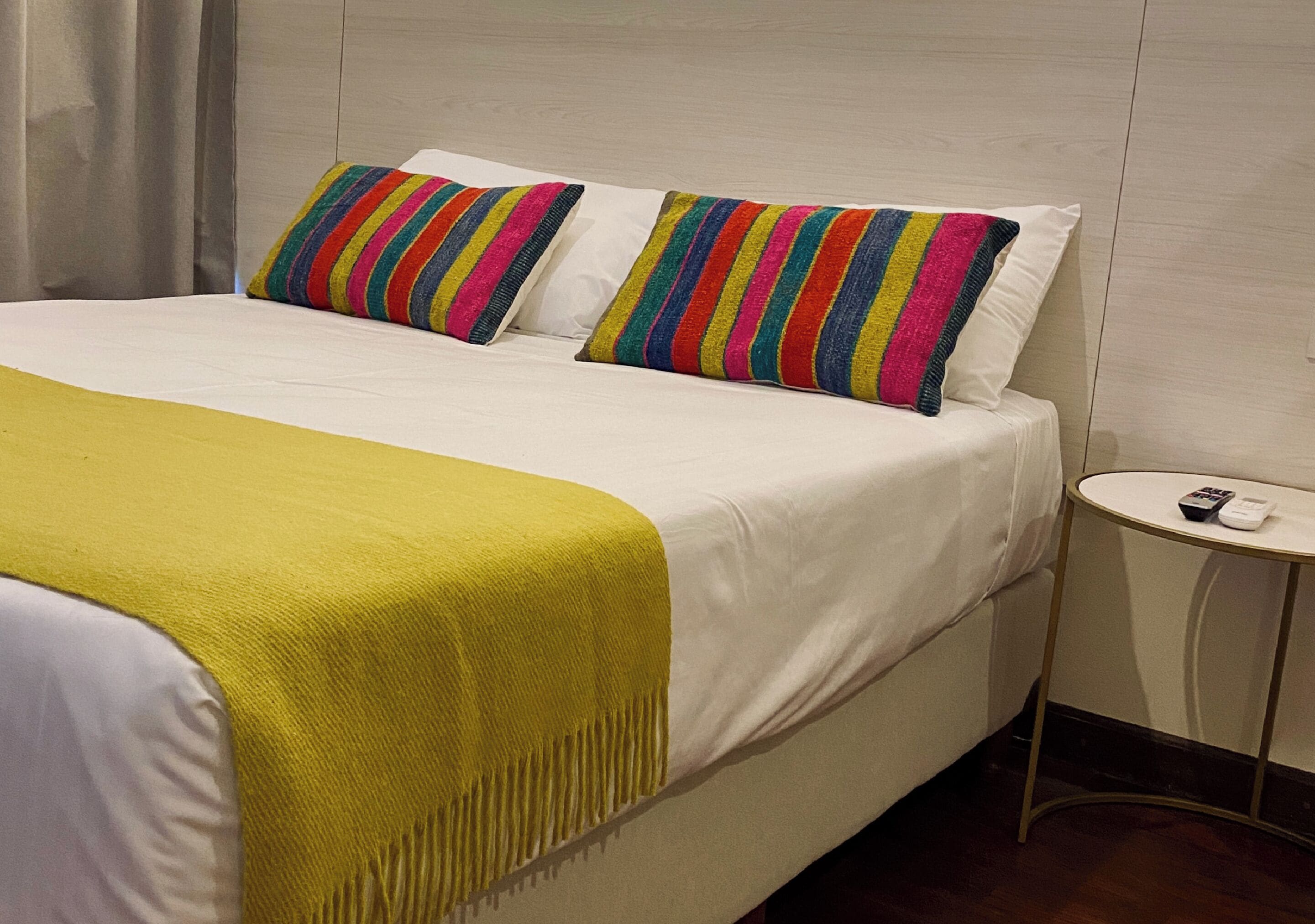 Una cama bien hecha con un plaid amarillo y dos coloridos cojines a rayas, una mesita de noche pequeña con un control remoto y un cabecero de madera.