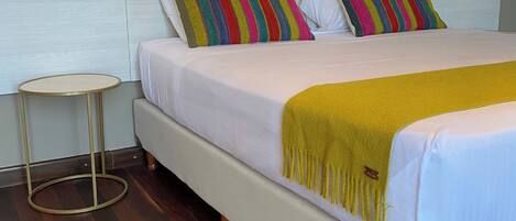 A bed with white bedding and a yellow throw blanket, two striped pillows, and a small round table with a gold frame.