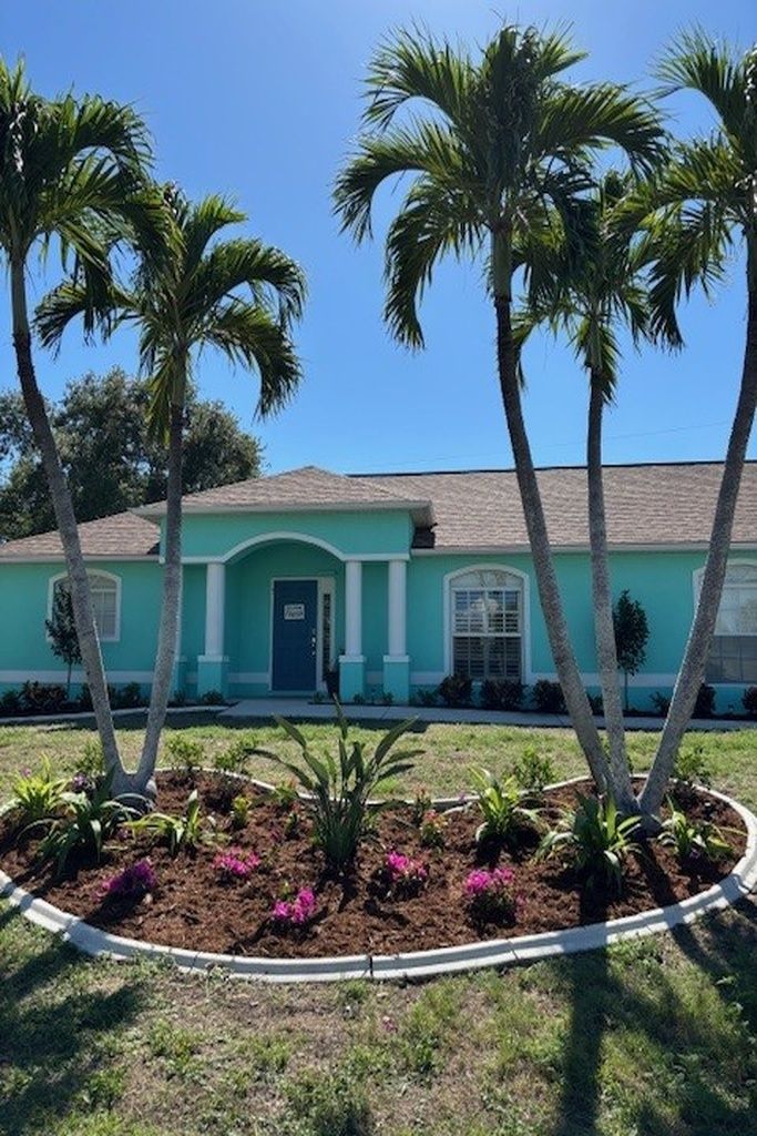 A single-story house with a turquoise facade, a dark blue door, and a small front yard with palm trees and flowering plants.