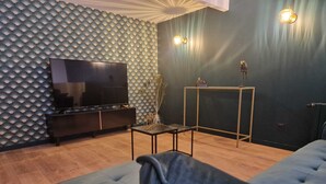 A modern living room with a large flat-screen TV, a dark blue sofa, a small coffee table, and a decorative wall with a fan pattern. - Gîte Bien-être Avec Jacuzzi & Sauna à la Campagne (Saint-Victurnien)