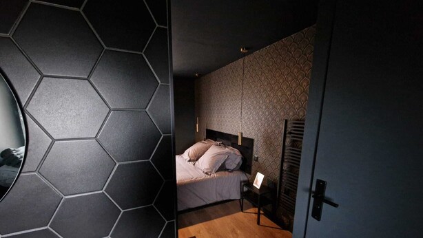 A modern bedroom with a hexagonal tile wall, a bed with a headboard, a bedside table, and a nightstand. - Gîte Bien-être Avec Jacuzzi & Sauna à la Campagne (Saint-Victurnien)