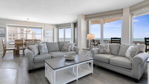 A spacious living room with a gray sectional sofa, a coffee table, and a dining area with wooden chairs and a table. Large windows provide a view of the ocean.