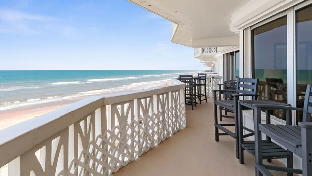 A balcony with a view of the beach, outdoor seating, and a white railing.