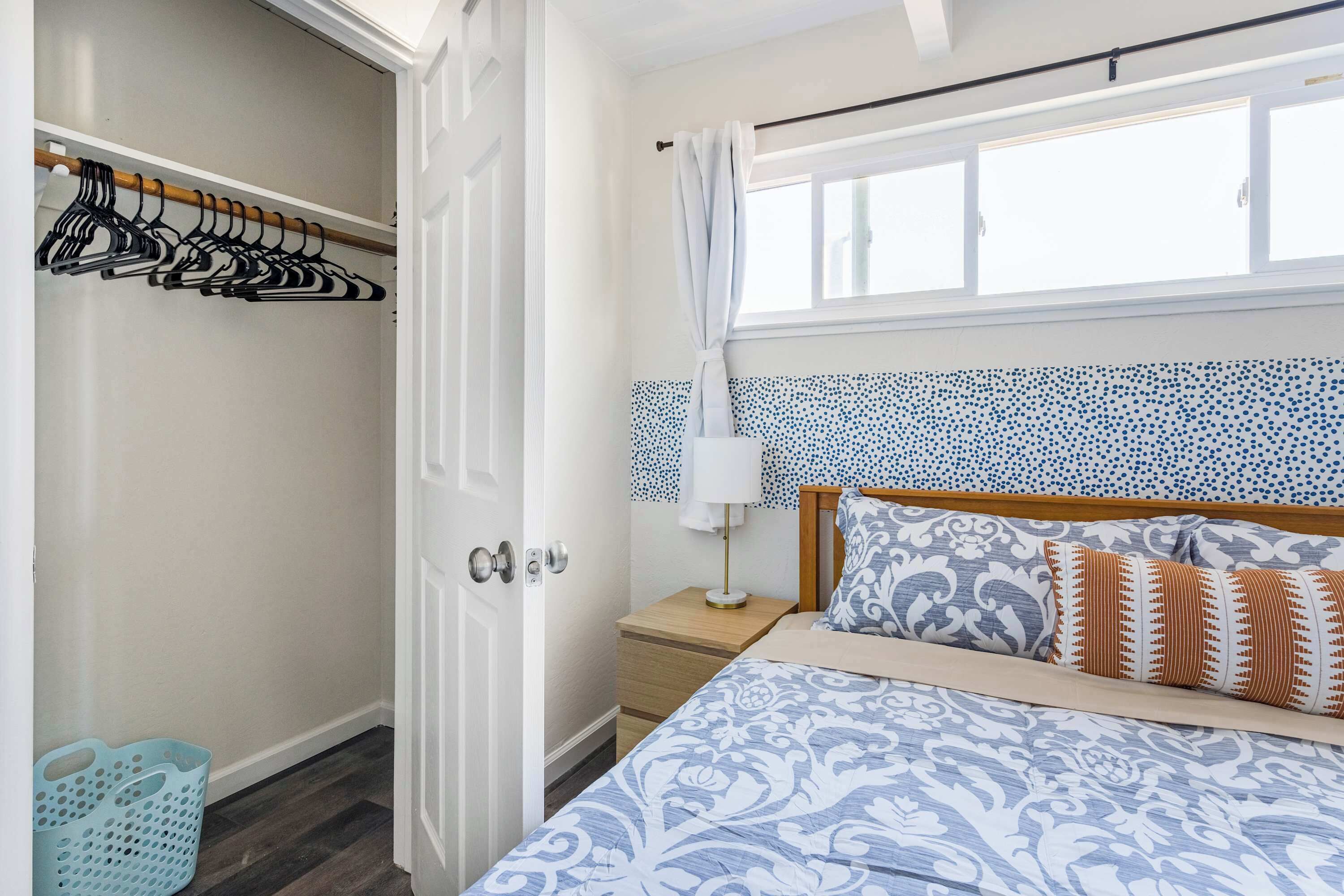 A bedroom with a bed, a nightstand, a lamp, a window with curtains, a closet with hangers, and a laundry basket.