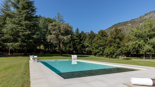 Une piscine rectangulaire à l’eau d’un bleu limpide, entourée d’une terrasse carrelée. On y trouve deux chaises longues blanches et un banc sur lequel est posée une serviette enroulée. La piscine est située dans un espace verdoyant, agrémenté d’arbres et avec une montagne en arrière-plan.