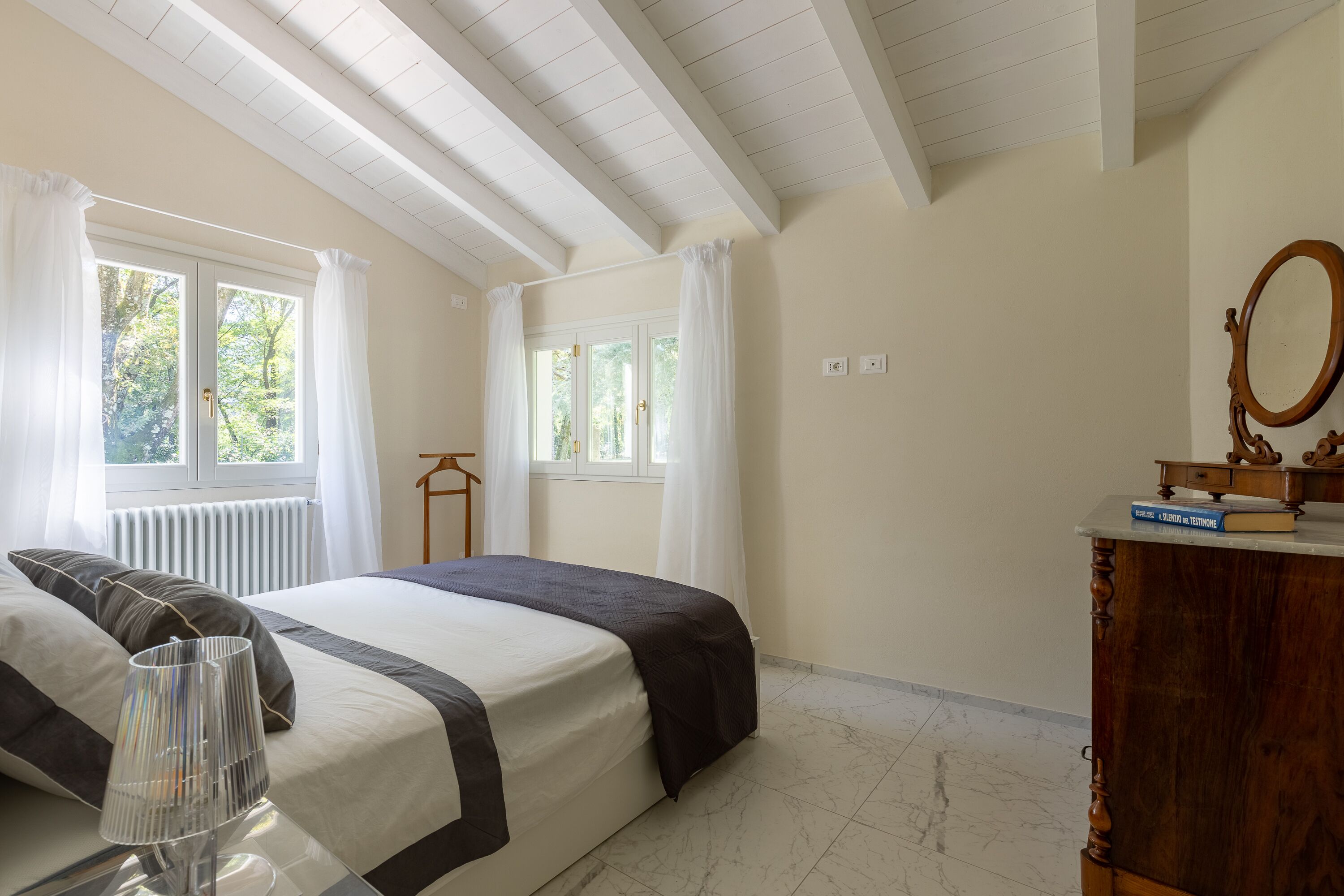 Un dormitorio con una cama grande, un aparador de madera, un espejo y dos ventanas con cortinas.