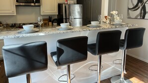 A modern kitchen with white cabinets, stainless steel appliances, and a marble countertop. There are three black bar stools and a white bowl on the counter. - THE UPTOWN BUNGALOW-COVERED PARKING-Walkable Historic Neighborhood-PET FRIENDLY! (Dallas)