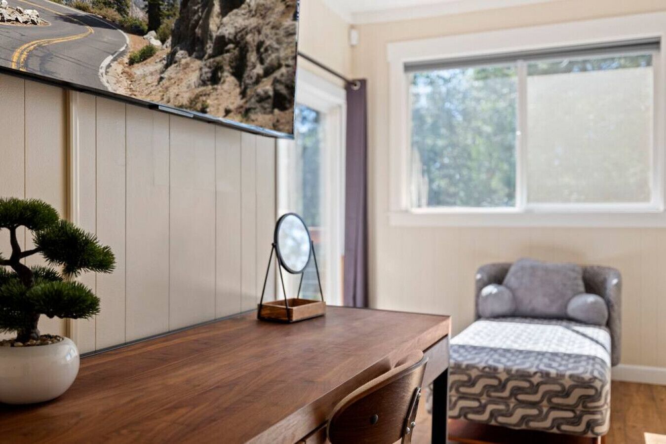 Une pièce avec un bureau en bois, une petite plante en pot, un miroir posé sur le bureau, une chaise grise avec des coussins et une fenêtre donnant sur des arbres.