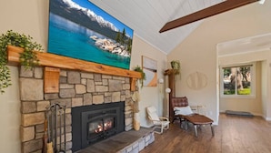 A living room with a stone fireplace, a large painting of a lake, and a view of the outdoors through a window. - Private Villa W/ Pool/spa, GameBarn, 4300 sq ft- Perfect for families and large groups (Twain Harte)