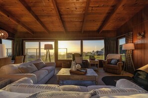 A cozy room with a wooden ceiling, large windows with curtains, a sofa, a chair, and a small table.