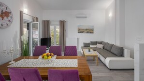 Uma sala de estar moderna com uma mesa de jantar de madeira, cadeiras roxas, um sofá cinzento e um quadro na parede.
