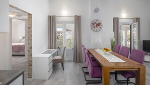 Uma sala de estar moderna com uma mesa de jantar de madeira, cadeiras roxas, um sofá branco, uma pequena escrivaninha e uma janela grande com cortinas.