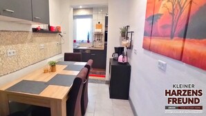 A modern kitchen with a stone backsplash, a dining area with a wooden table and black mats, and a living area with a large abstract painting.