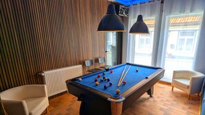 A pool table in a room with wooden paneling, two chairs, and a window with a view of buildings.