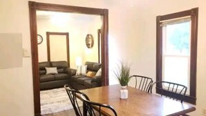 Un comedor con una mesa y sillas de madera, una planta en maceta y vistas de una sala de estar con un sofá y una lámpara.