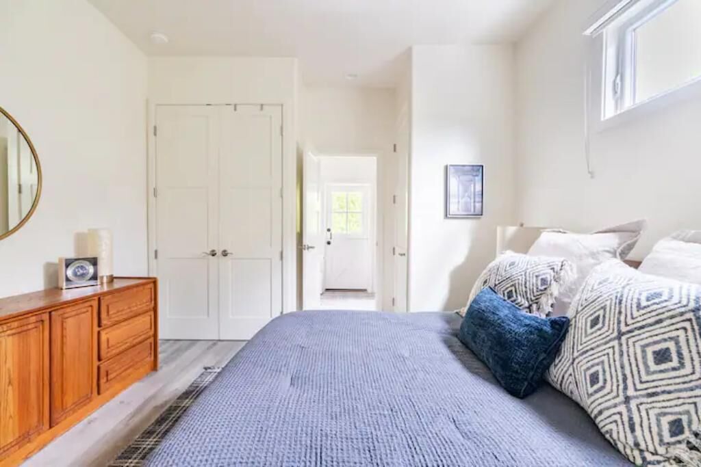 A bedroom with a large bed, a wooden dresser, a mirror, and a door leading to another room.