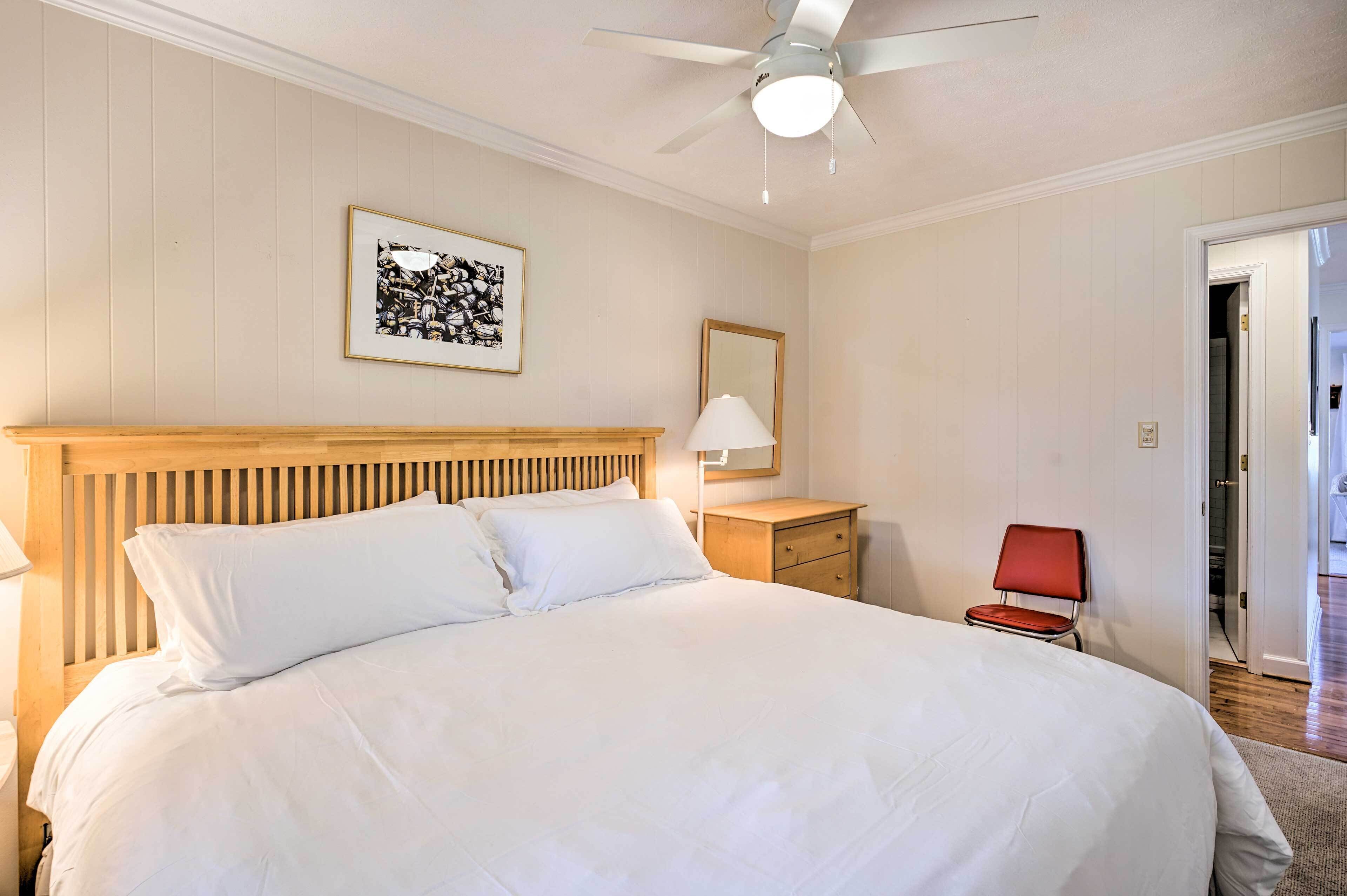 A neatly made bed with white linens, a wooden headboard, a ceiling fan, a framed black and white photo, a bedside lamp, a wooden nightstand, and a red chair.