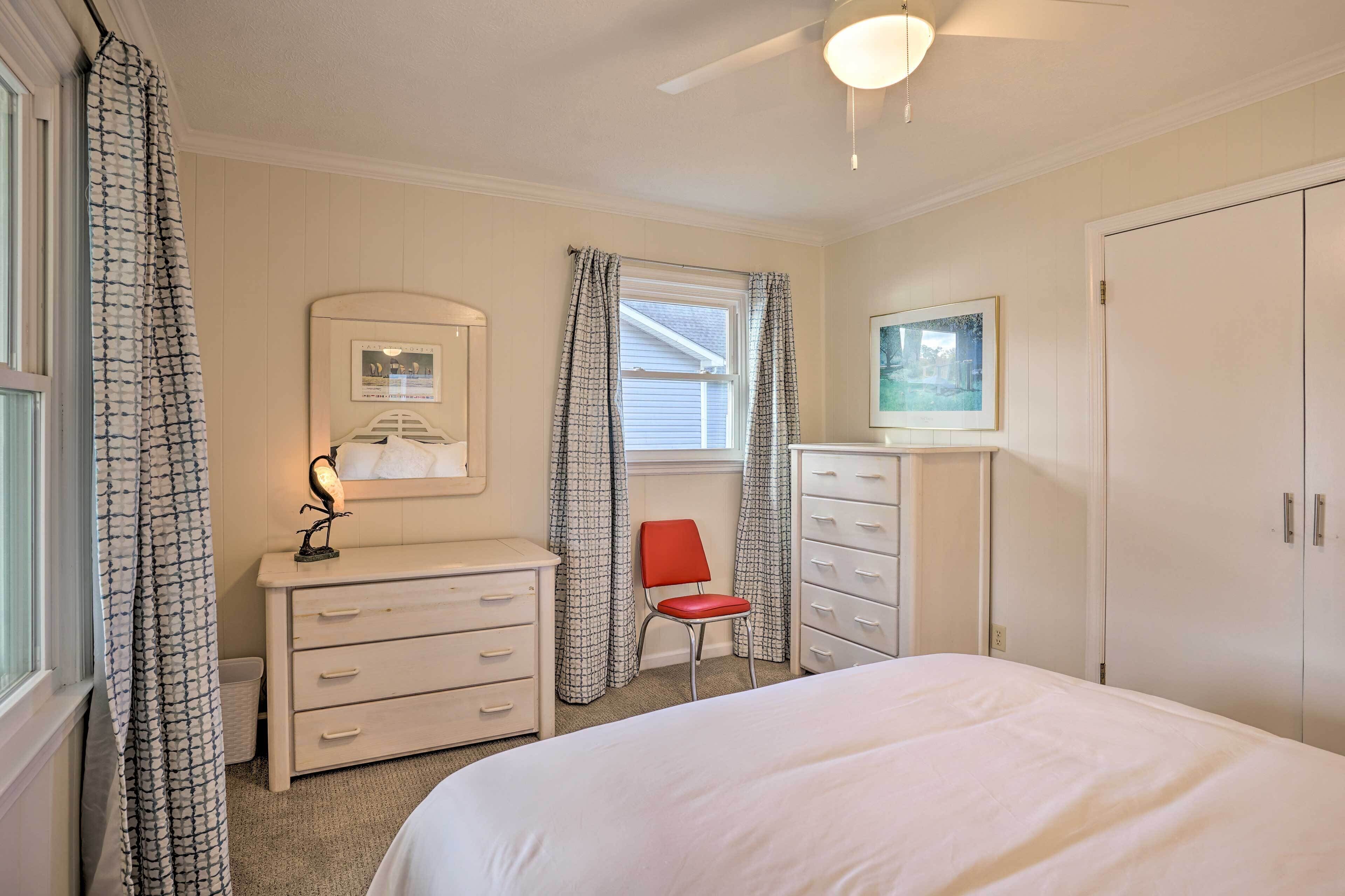 Een slaapkamer met een bed, een dressoir, een stoel, een bureau, een raam met gordijnen en een plafondventilator.