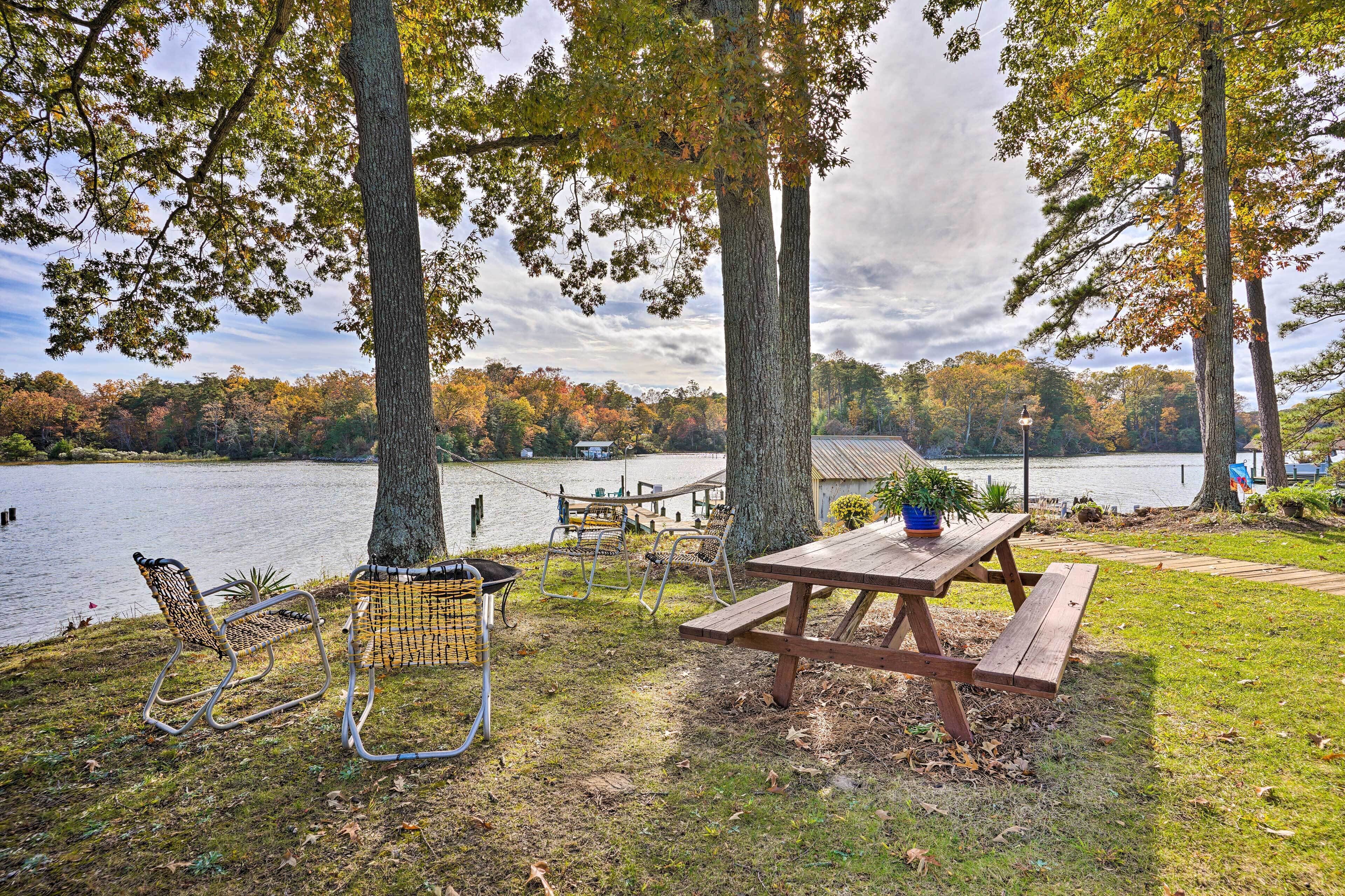 Ein Picknickplatz am See mit einem Holztisch und Metallstühlen, umgeben von Bäumen und Seeblick.