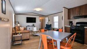 Un espacio habitable compacto con kitchenette, una zona de comedor con sillas naranjas, un sofá y un televisor montado en la pared.