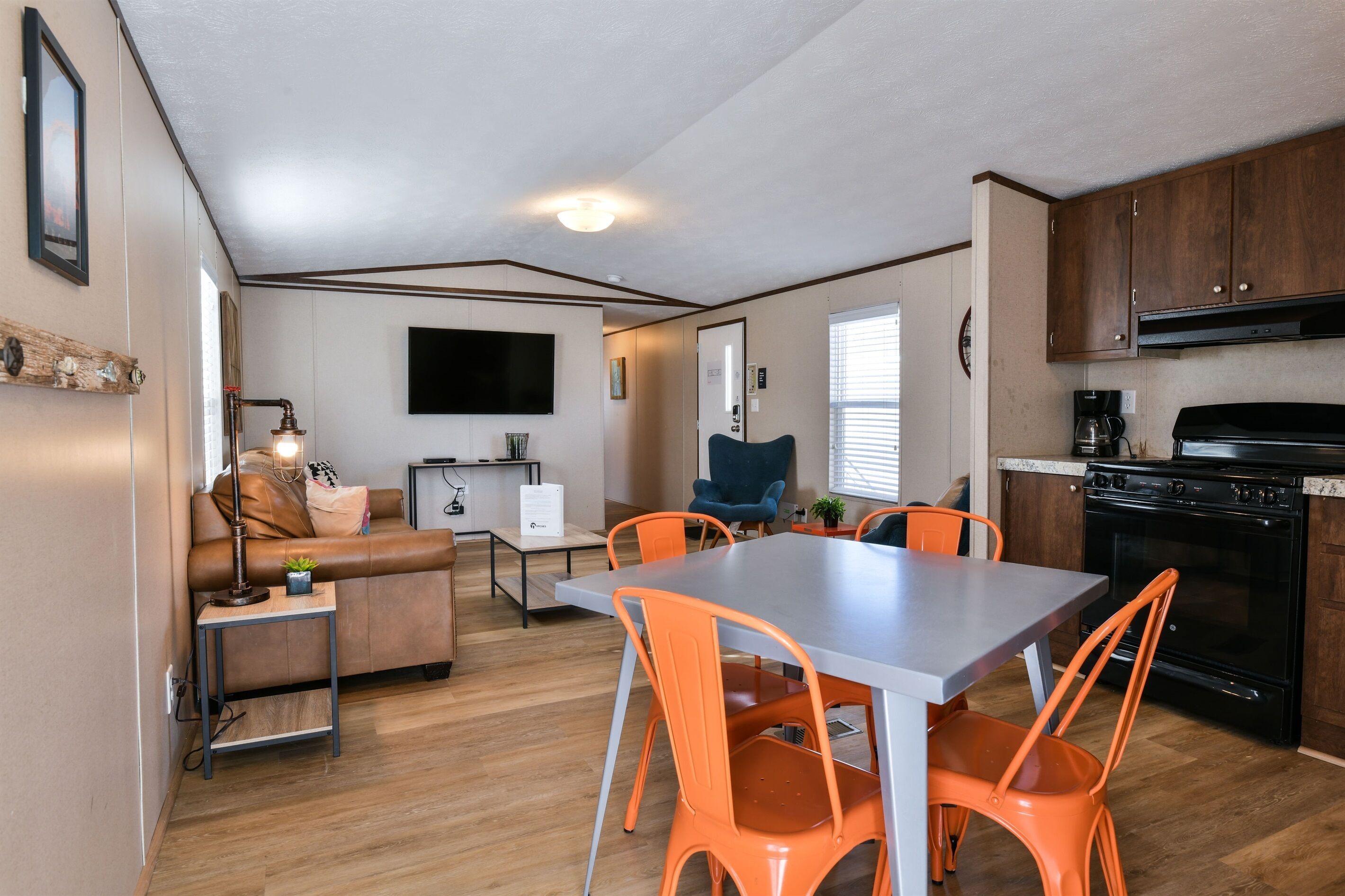A compact living space with a kitchenette, a dining area with orange chairs, a sofa, and a TV mounted on the wall.