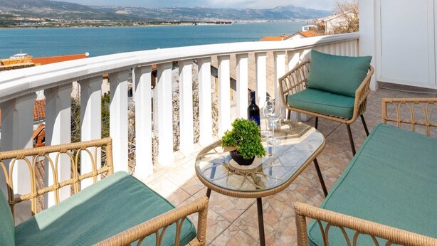 A balcony with wicker furniture, a glass table, and a view of the sea and mountains.