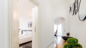 A modern hotel room with a bed, a wooden side table with a potted plant, and a hallway with a white door leading to another room.