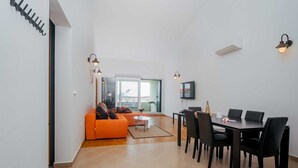A modern living room with a sofa, dining table, and chairs. There is a TV mounted on the wall and a window providing a view of the outdoors. - Apartment Jagoda near the beach (Okrug Gornji)