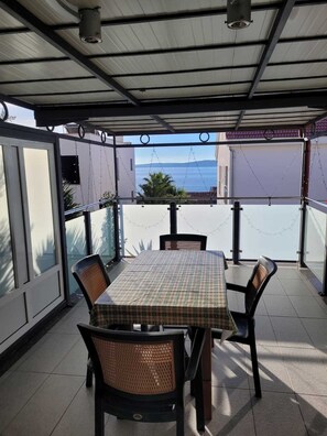 Eine überdachte Terrasse mit Tisch und Stühlen, Blick aufs Meer und ein Gebäude im Hintergrund.