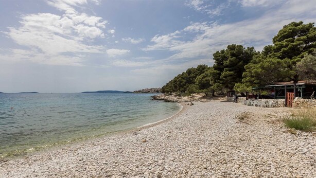 A pebble beach with clear water, a wooden structure, and trees.