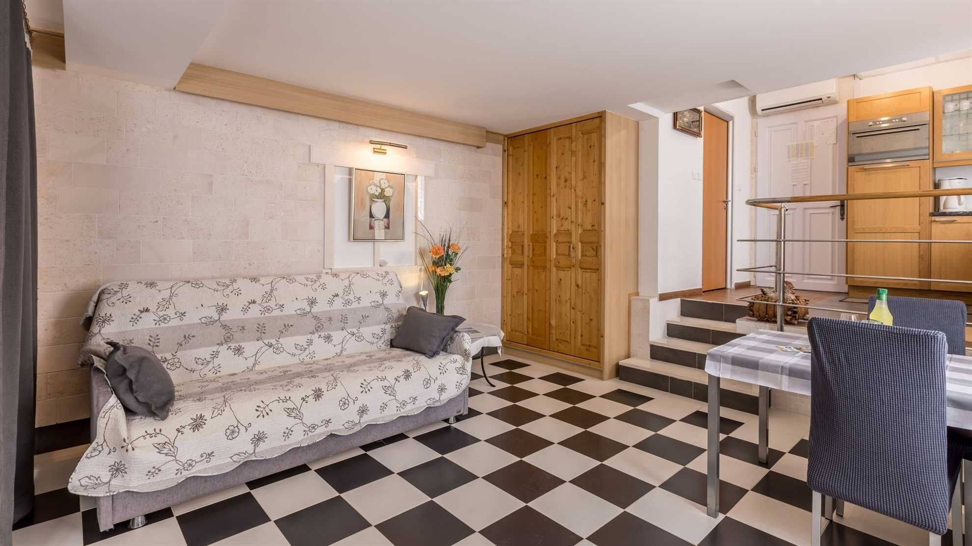 A living room with a checkered floor, a sofa, a dining table with chairs, and a kitchen area with wooden cabinets and an oven.