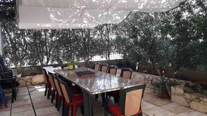 An outdoor patio with a long table and chairs, surrounded by potted plants and trees. - Studio - apartment Katarina - EOS CROATIA (Okrug Gornji)