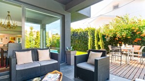 Une terrasse extérieure avec des meubles en osier, une porte vitrée donnant sur une piscine et une vue sur la verdure.
