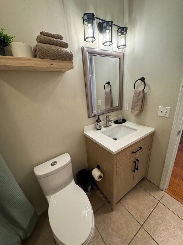 Baño con inodoro, lavabo y espejo. Hay toallas en un estante y un cubo de basura.