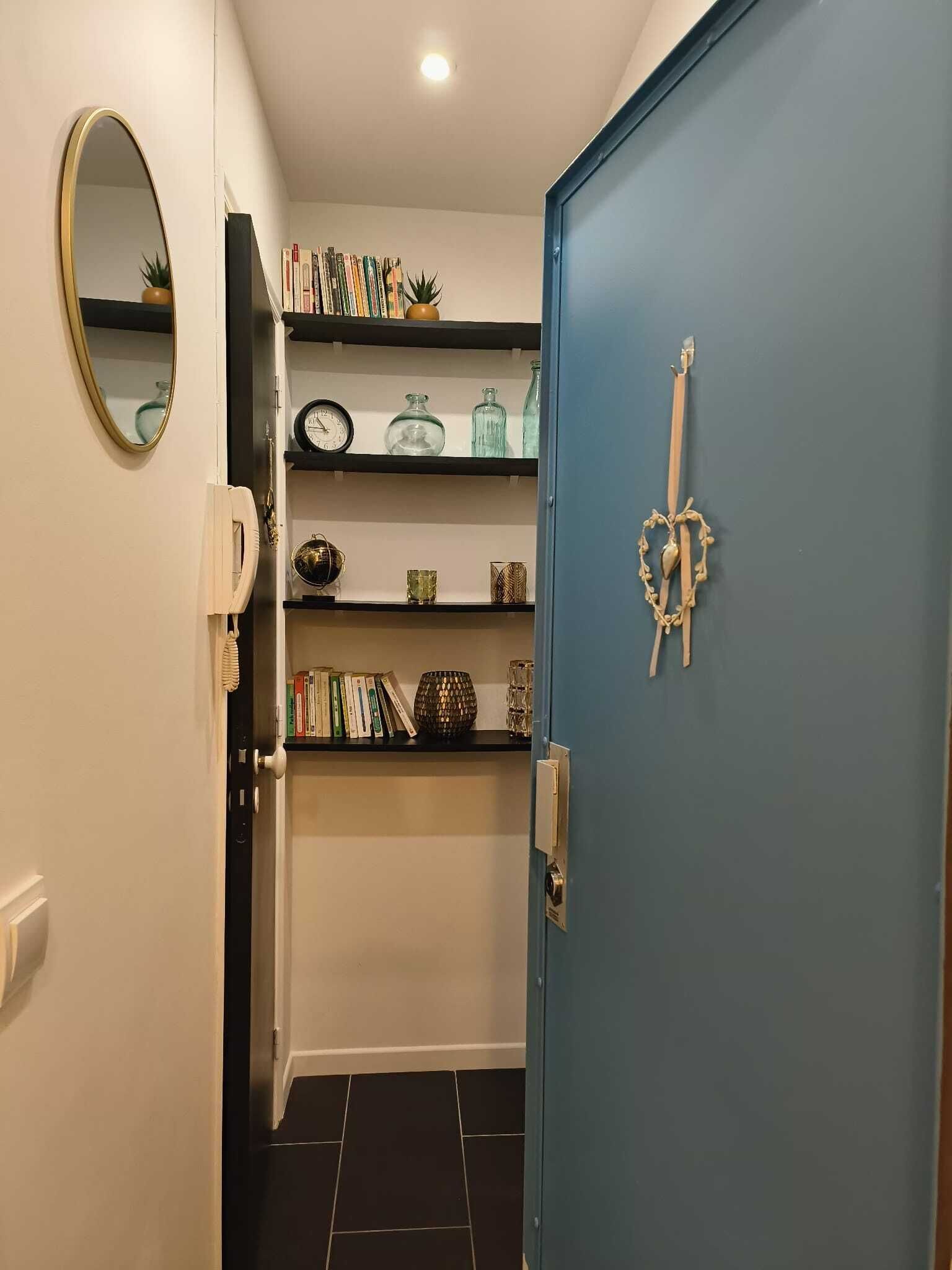 A hallway with a blue door, a mirror, and shelves with books and decorative items.