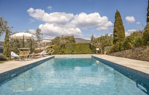 Una piscina con sillas de descanso y sombrillas, rodeada de vegetación y un muro de piedra.