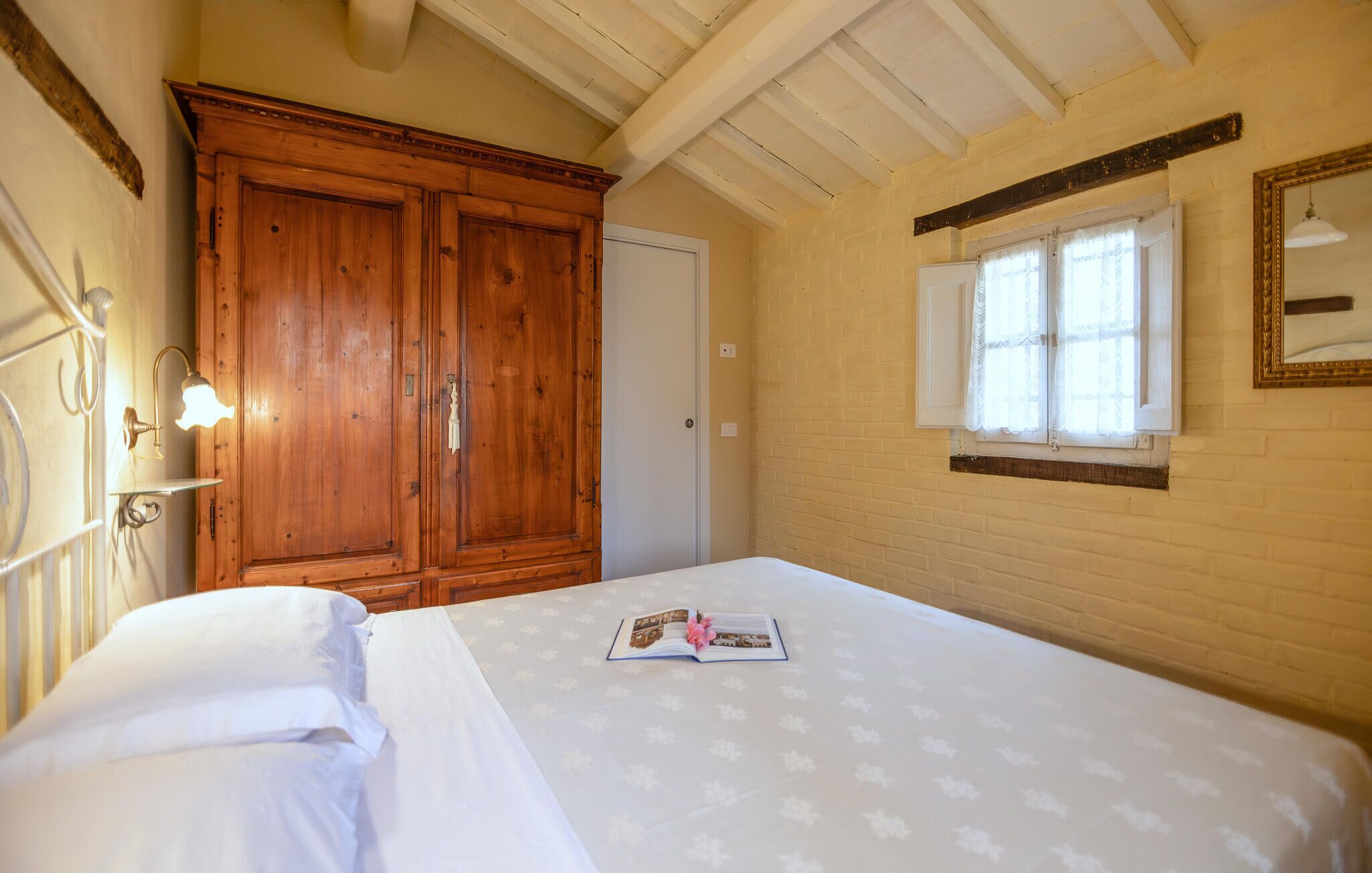 Een slaapkamer met een grote houten garderobe, een bed met een witte dekbedovertrek, een raam met kantgordijnen, en een spiegel aan de muur.