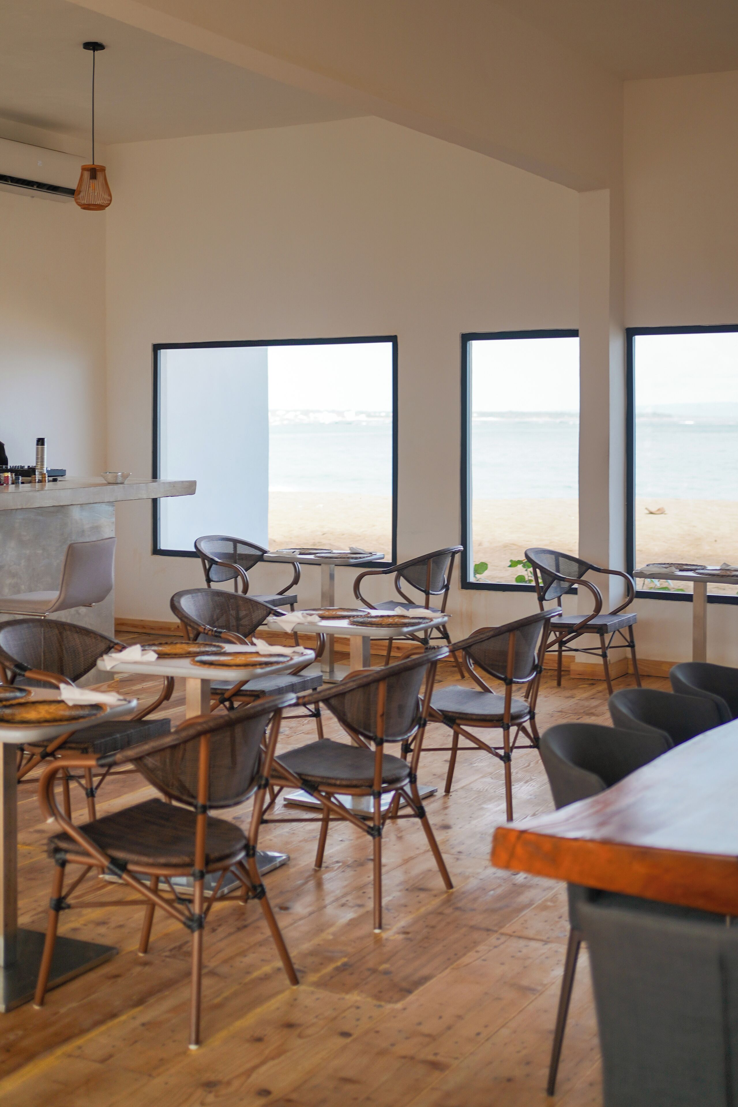 Um interior moderno com soalho em madeira, cadeiras de estrutura metálica e amplas janelas com vista para uma praia.