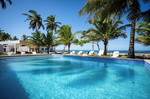 Piscina azul límpida com cadeiras de banho brancas e palmeiras, emoldurada por um edifício moderno e vista para o oceano.